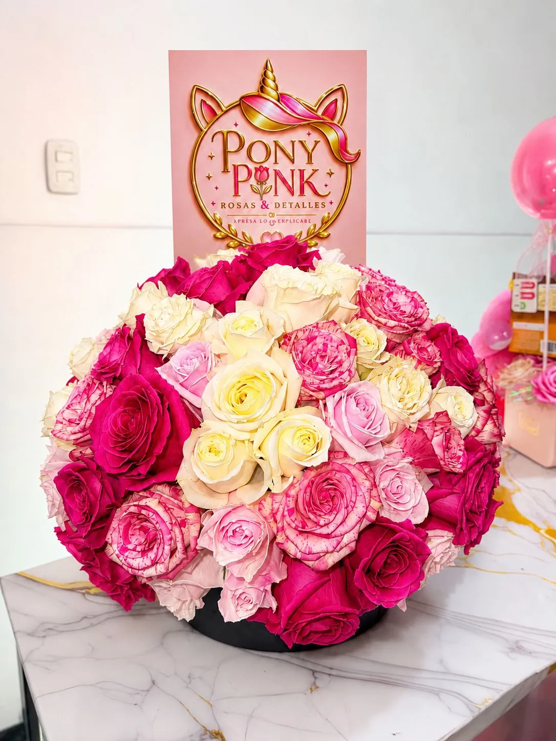 Arreglo circular de rosas fucsia, rosadas y crema en una base negra, con una tarjeta rosa de PonyPink detrás.