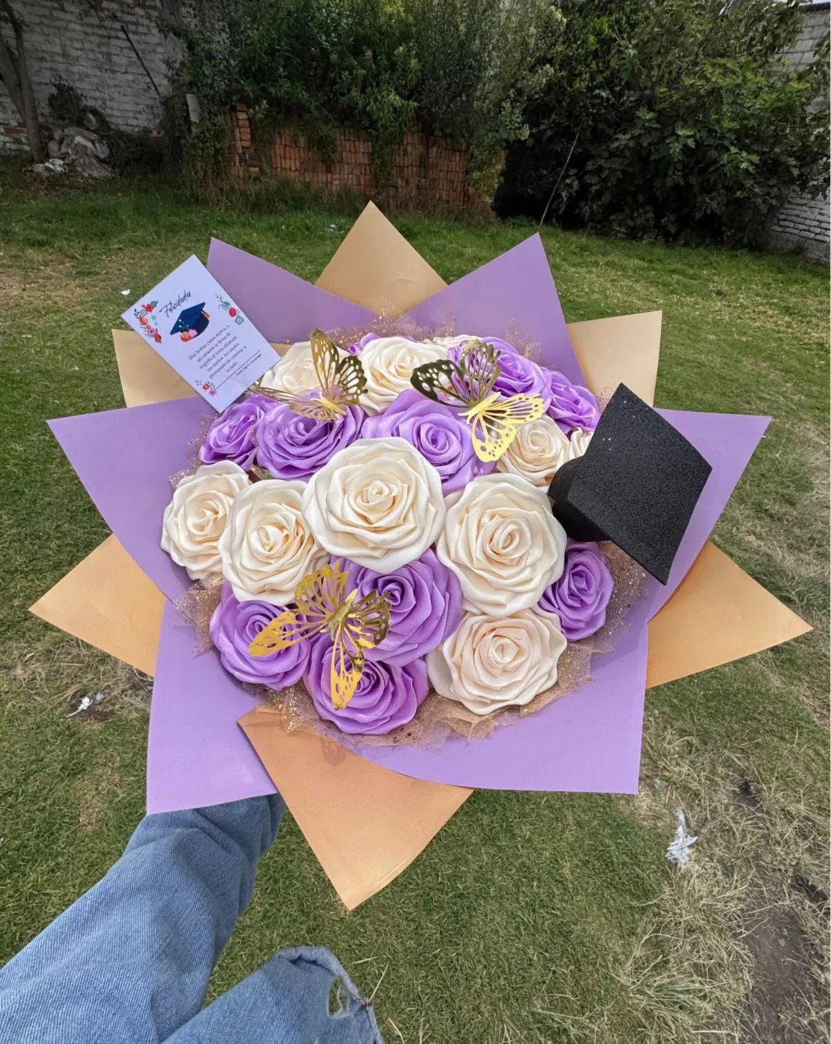 Ramo de rosas de cinta lilas y crema con mariposas doradas, envoltura lila y durazno en picos, un birrete negro y una tarjeta de graduación.