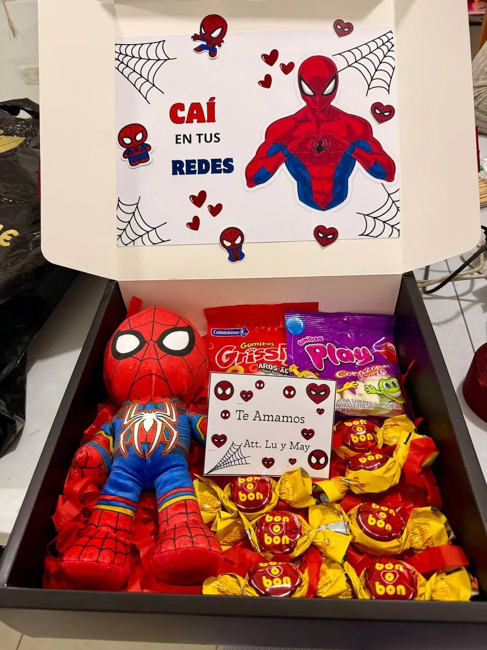 Caja abierta con un peluche de superhéroe, golosinas y una tapa decorada con una dedicatoria.