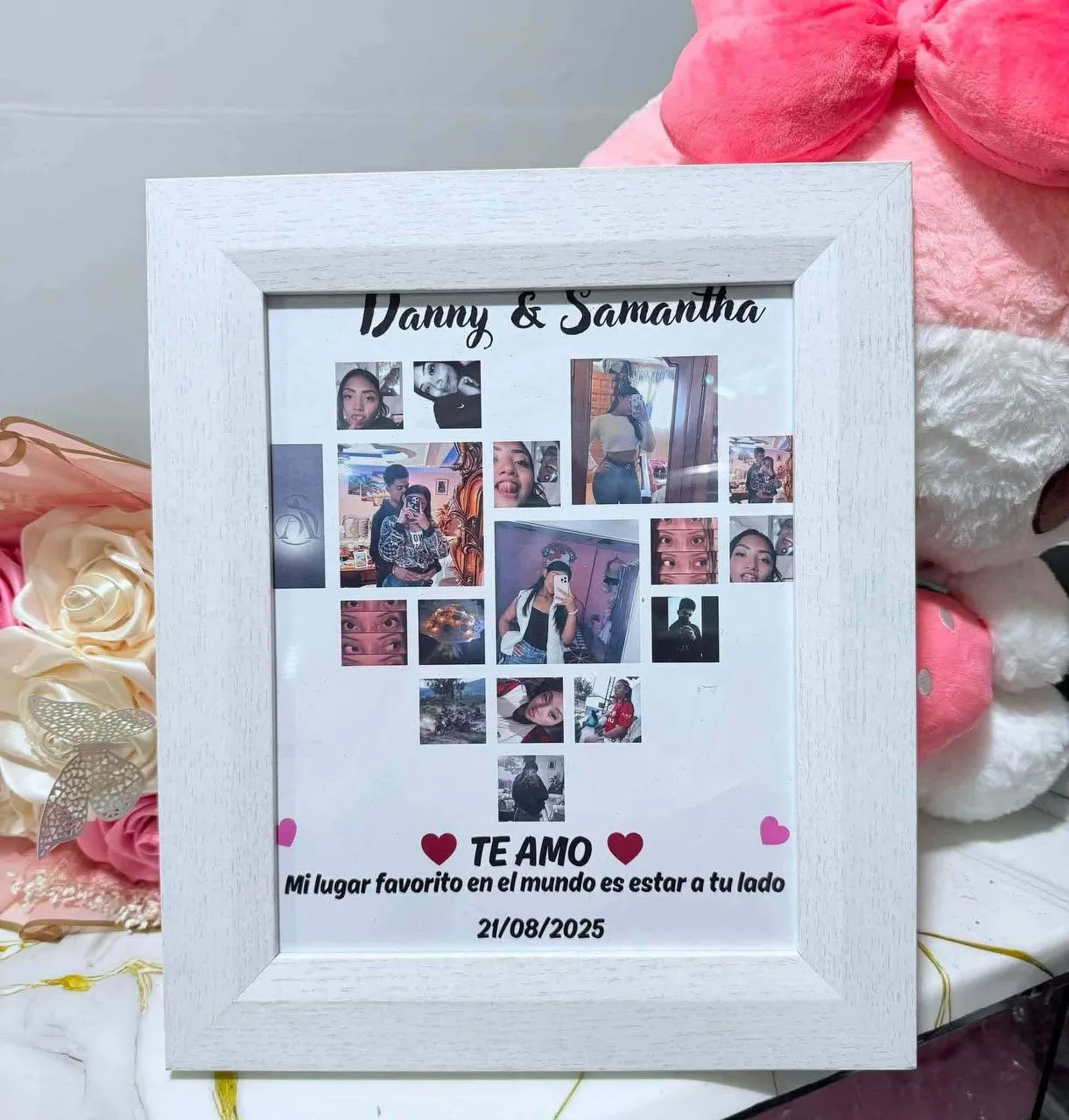 Cuadro blanco con un collage de fotos en forma de corazón y una dedicatoria, junto a peluches y flores.