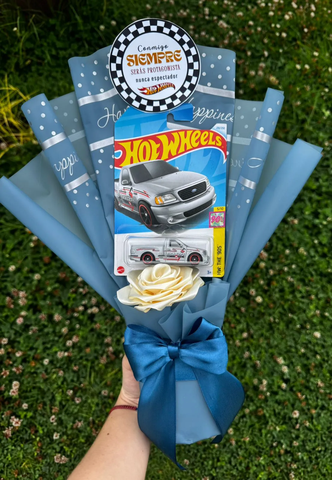 Ramo azul con un carro de juguete en su blíster, una rosa de cinta blanca, lazo azul y un cartel redondo con una dedicatoria.