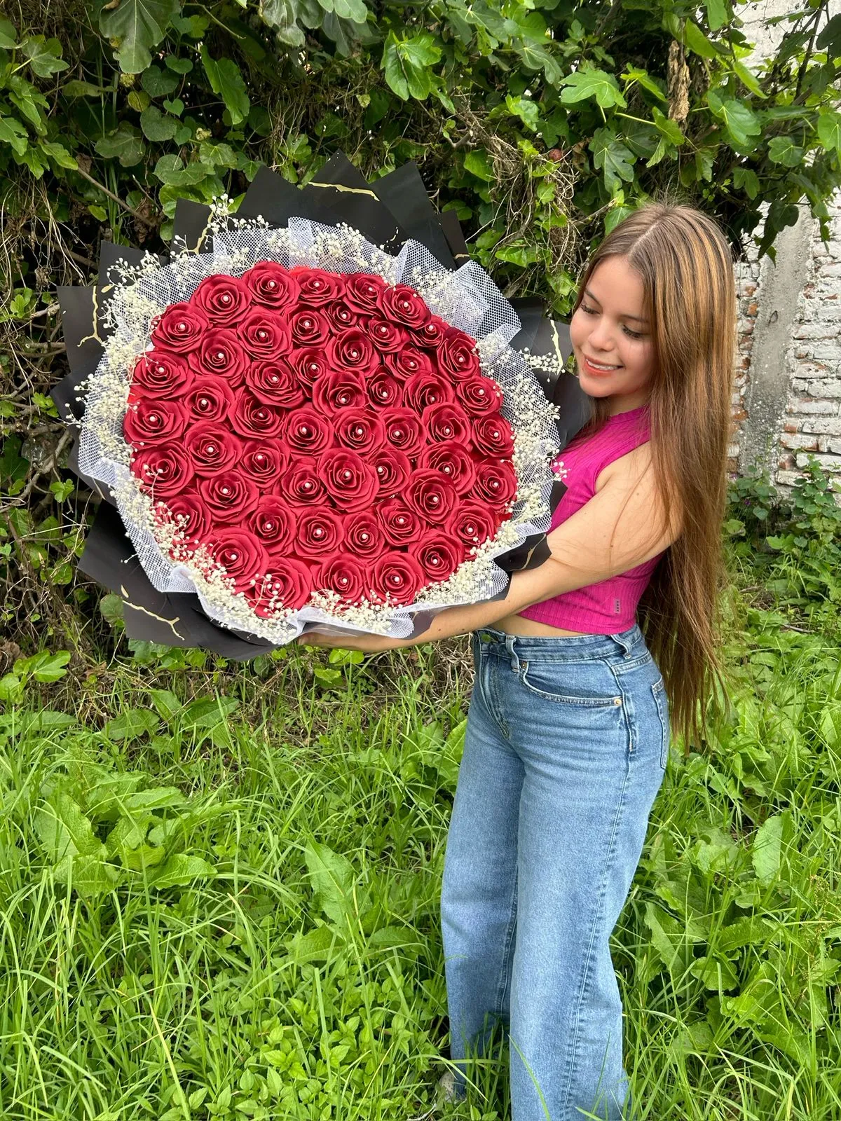 Una mujer sostiene un gran ramo de rosas rojas con envoltura negra y flores blancas frente a un fondo de plantas.
